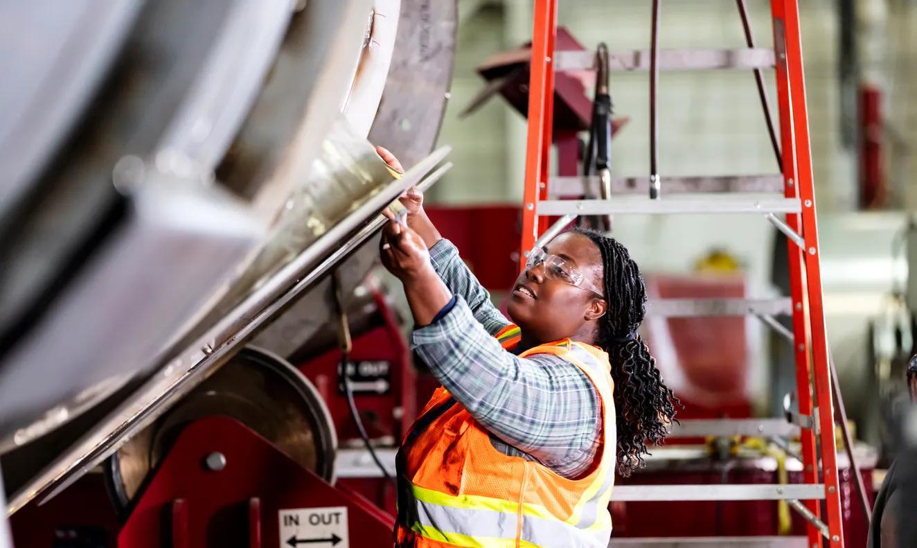 A woman wearing a safety vest and protective goggles works in a metal fabrication shop.  