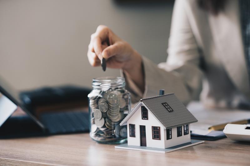 Hand of investor putting coin in jar next to wooden house